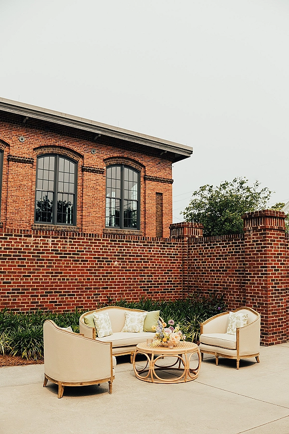 Lounge seating area with wedding lounge furniture, outdoor sofa and chairs around a rattan coffee table with floral centerpiece on a brick patio