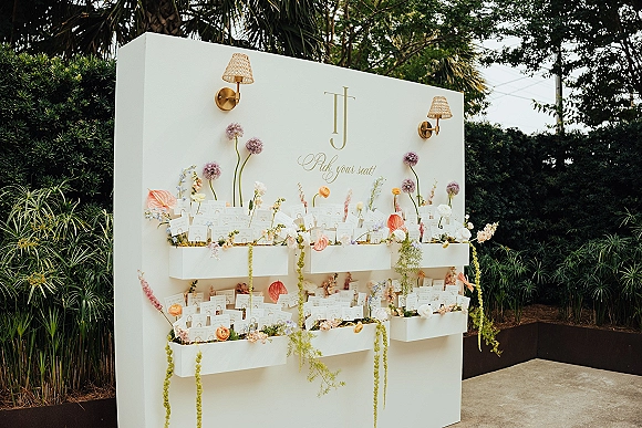 Wedding seating chart with escort card display on a white wall, gold lettering and monogram, floral arrangements and hanging greenery in a garden patio setting