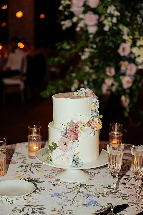 Wedding cake with white buttercream, two-tier wedding cake topped with fresh flowers on a stand, bistro lights and greenery wall behind