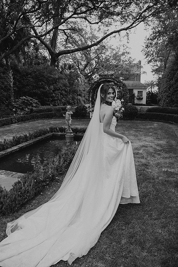 Bridal portrait of a bride in long veil holding a bouquet, looking back on a garden lawn near a fountain and statue with archway beyond