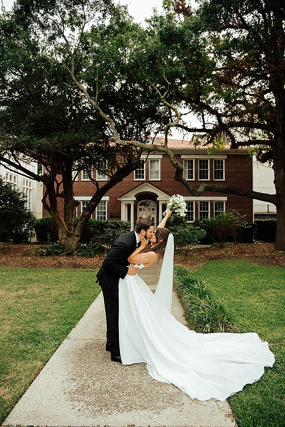 Wedding kiss portrait of bride and groom kiss in a romantic dip, bouquet raised, veil and long train flowing by a brick building walkway