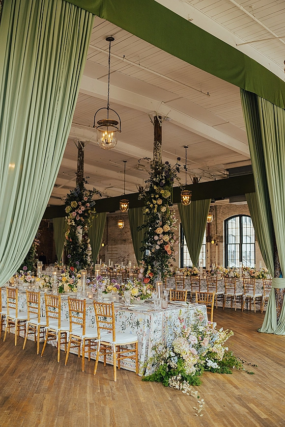 Reception tablescape with long banquet table wedding details, pastel floral centerpieces and tall cylinder candles beneath green draping in a brick loft