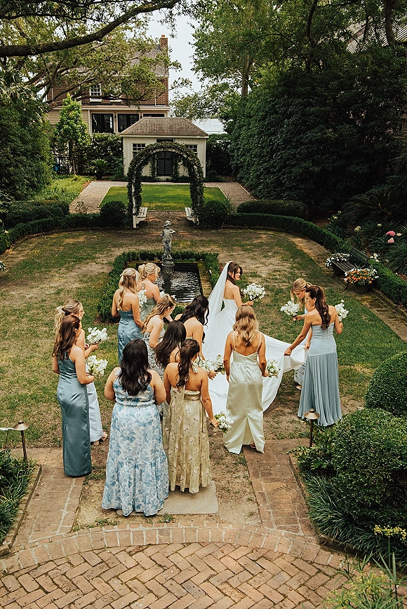 Bridal party photo of bride with bridesmaids holding her veil and bouquets in a circle on a brick garden path by a fountain