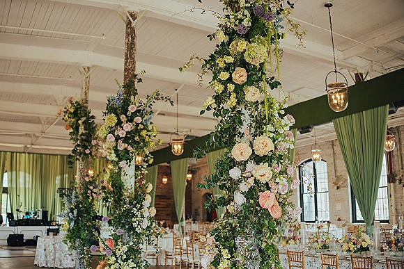 Reception floral installations with hanging wedding flowers above banquet tables, greenery-wrapped columns, pastel centerpieces in an industrial loft