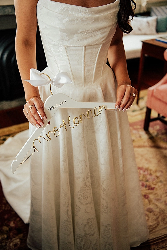 Wedding dress detail on a custom wedding hanger with white ribbon bow, engagement ring and manicure in a getting ready room