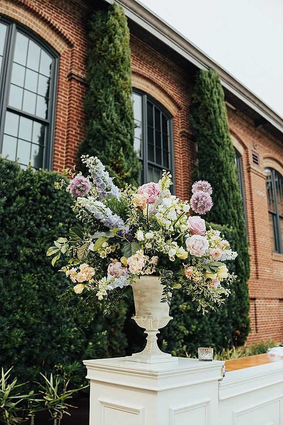 Wedding floral arrangement in an urn vase centerpiece with lush greenery atop a white pedestal at a brick venue with arched windows