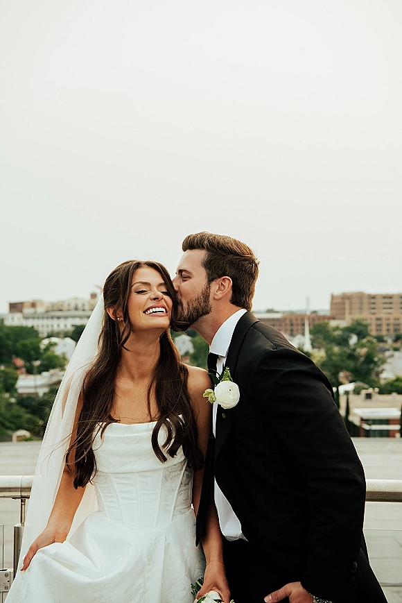 Wedding couple portrait of groom kissing bride’s cheek as she laughs, her long veil flowing on a rooftop terrace with city skyline