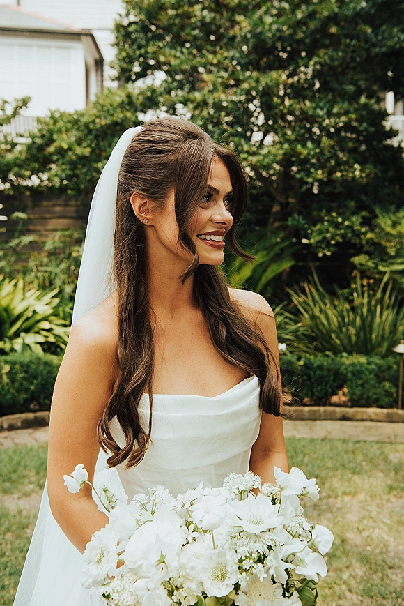 Bridal portrait of a smiling bride holding bouquet in a strapless gown and veil, looking to the side in a lush garden lawn setting