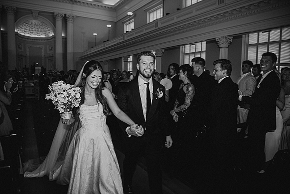 Wedding recessional as bride and groom walk the aisle holding hands, bride holding bouquet, guests applauding in a grand church interior