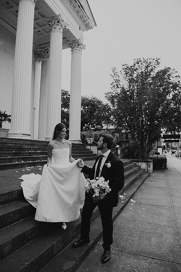 Couple portrait of bride and groom on steps, she holds her dress hem while he holds a white bouquet on stone courthouse stairs with columns