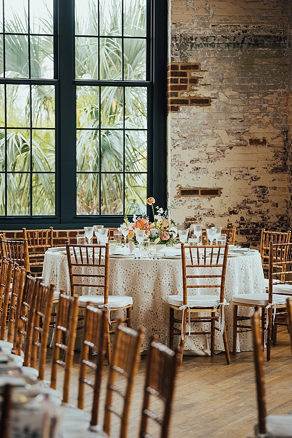 Reception tablescape with a round wedding reception table dressed in lace tablecloth, pastel florals, bud vases, and glass goblets by brick wall windows