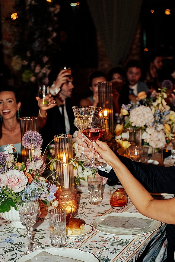 Wedding toast with raised wine glasses over a candlelit reception table, pastel floral centerpieces and dimly lit guests in back
