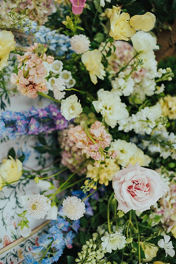 Wedding flowers in a floral patterned box, featuring yellow roses, a pink rose, blue hydrangea, white blooms and greenery against dark foliage background