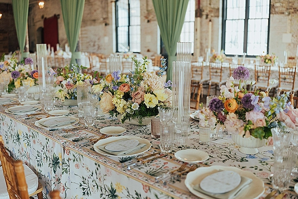 Reception tablescape with wedding table decor, colorful floral centerpieces, gold flatware, crystal glassware, and taper candles in a brick loft room
