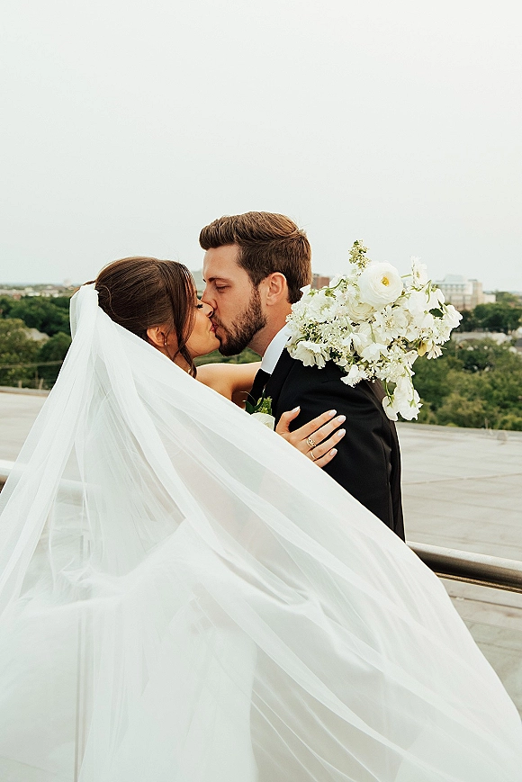 Wedding kiss portrait of bride and groom kissing as her long veil blows in wind, holding a white bouquet on a rooftop terrace with city skyline