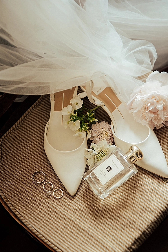 Bridal accessories flatlay with ivory wedding heels, rings and perfume bottle on a striped chair, framed by veil and blush flowers