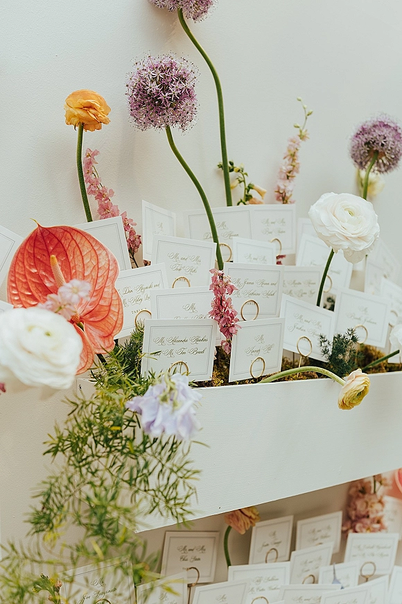 Wedding escort cards on a white shelf display, clipped with gold paper clips and framed by ranunculus, allium, anthurium, and moss against a white wall