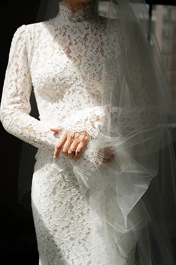 Lace wedding dress with high neck bodice and long sleeves, bride’s hands showing engagement ring and manicure in window light near brick wall