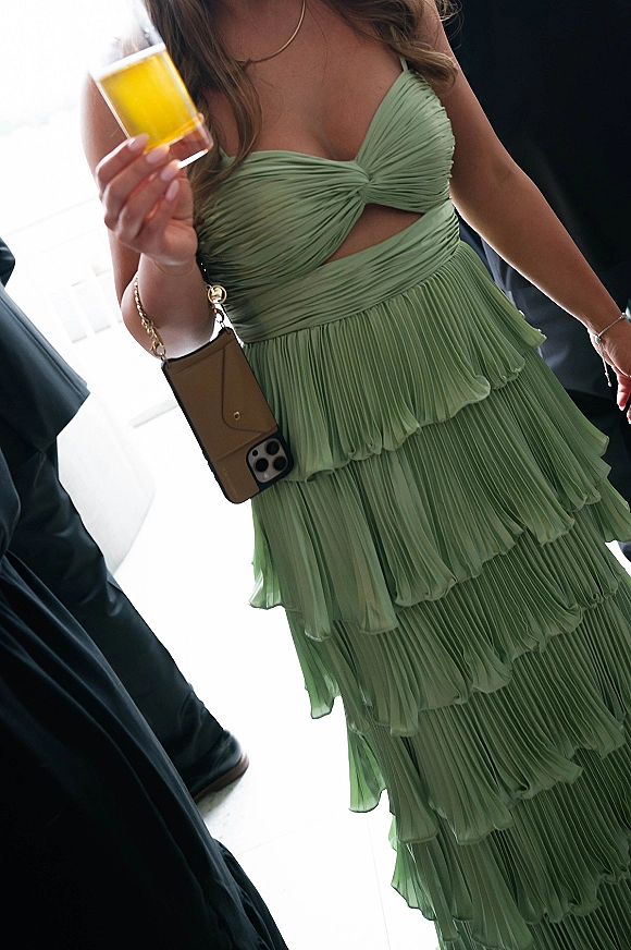 Wedding guest dress in sage green with pleated ruffles and a cutout bodice, holding a cocktail glass in a doorway with dark suits behind