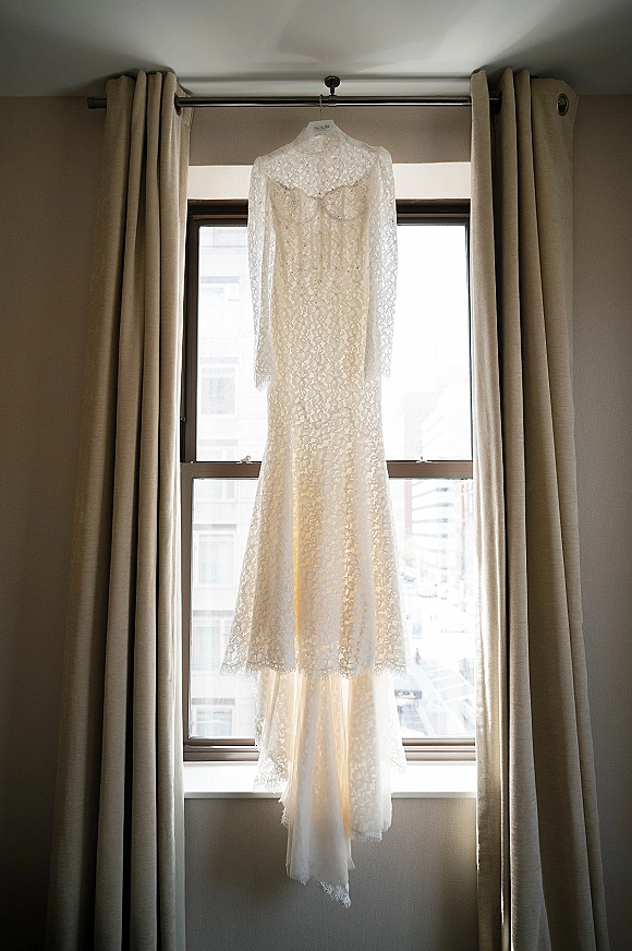 Wedding dress with lace wedding dress details, long sleeves and high neck, hanging on a hanger by a bright window with city buildings beyond