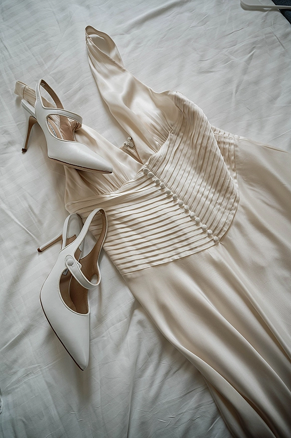 Wedding dress detail with satin wedding dress pleated bodice and fabric-covered buttons beside white slingback heels on white bed sheets