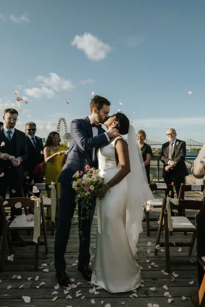 Wedding kiss as rose petals fall on a wooden dock aisle, bride’s veil flowing beside groom in bow tie with skyline and ferris wheel beyond