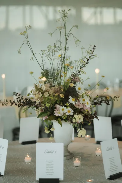 Wedding centerpiece with wildflower floral arrangement in a white ceramic vase, surrounded by votive candles and table numbers in a reception tent