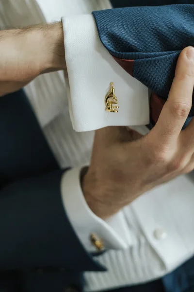 Groom cufflinks in gold on a white French cuff shirt as he adjusts his sleeve, with suit jacket and necktie visible in close-up