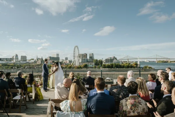 Wedding ceremony on a rooftop deck with bride and groom at a circle floral arch, guests seated, city skyline, river and ferris wheel behind