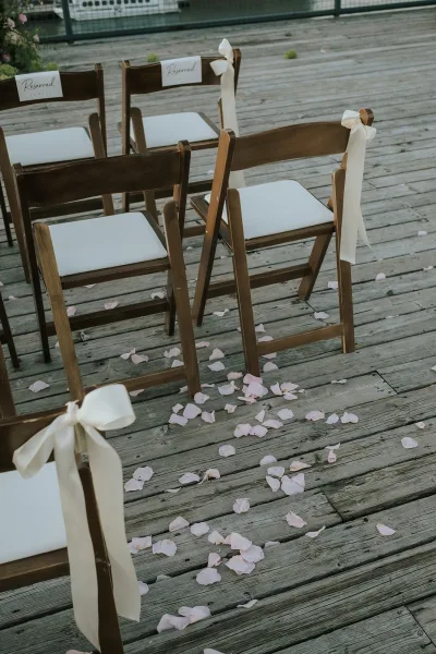 Ceremony aisle decor with wedding aisle rose petals scattered by white ribbon bows on wood folding chairs on a wooden deck with railing