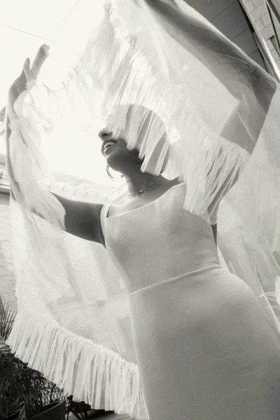 Bridal portrait of a bride holding veil, smiling and looking up in a sleeveless gown with necklace and earrings outdoors by a fence