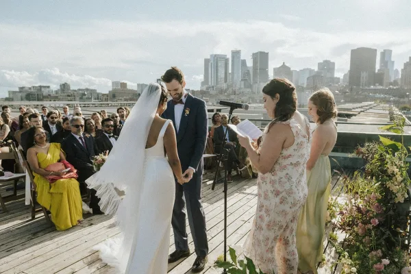Wedding vows as bride in a long veil and groom in a navy suit hold hands at an outdoor rooftop ceremony with city skyline backdrop