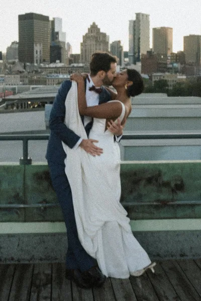 Wedding kiss portrait of bride and groom kissing in a dip on a rooftop deck, her veil flowing, with city skyline behind them
