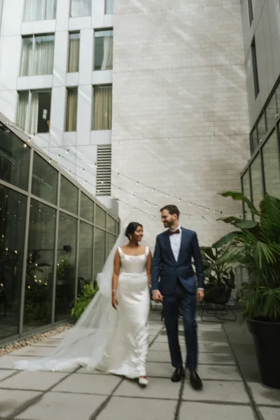 Couple portrait of newlyweds holding hands, bride in satin dress and long veil with groom in bow tie under string lights in a city courtyard
