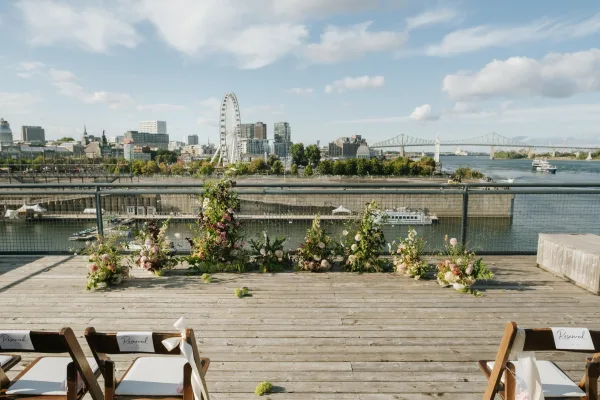 Ceremony setup with outdoor wedding ceremony aisle flowers lining a wooden deck, wooden chairs and reserved signs, river skyline beyond