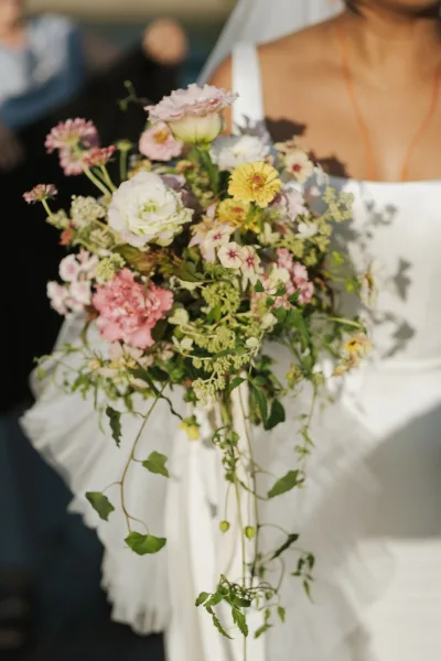 Bridal bouquet with wildflower bridal bouquet blooms and trailing greenery, tied with ribbon in sunlight as the bride holds it in a white dress