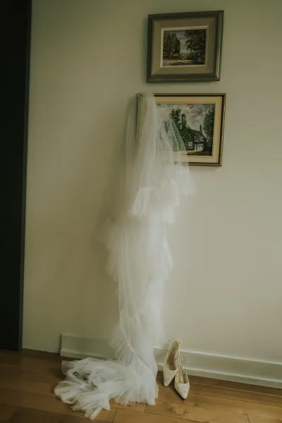 Wedding dress hanging on a wall hook with a bridal veil draped beside it, lace pointed-toe heels on the wood floor below