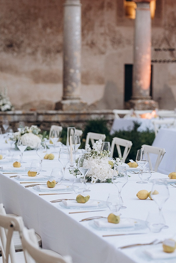 Reception tablescape with long banquet table, white floral centerpieces and lemons, gold flatware and place cards in a stone courtyard patio with columns