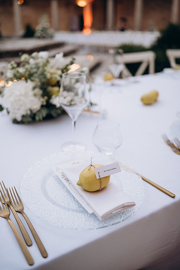 Reception tablescape with wedding place setting, glass charger, gold flatware, and lemon place card tied with twine in a stone courtyard lit by warm lights