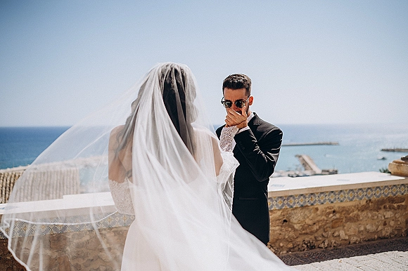 Wedding first look as bride walks up behind groom for an emotional reaction, long veil blowing on a coastal terrace by the ocean