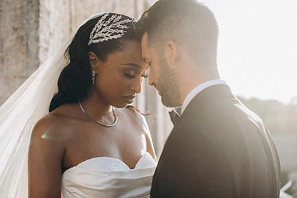 Wedding couple portrait of bride and groom close up touching foreheads, bride in veil and tiara, groom in tuxedo by a stone wall outdoors