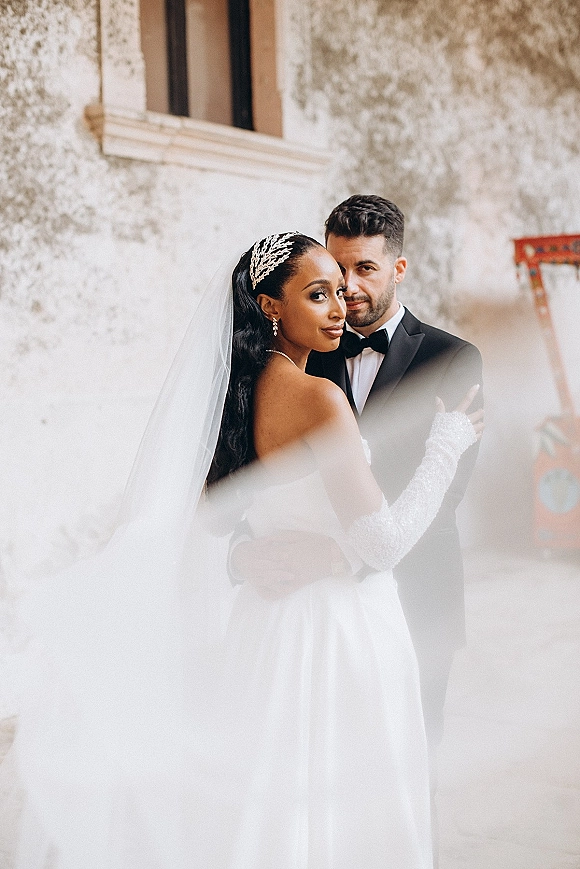 Couple portrait of bride and groom embrace, bride looking over shoulder in long veil against a textured stucco wall by a window