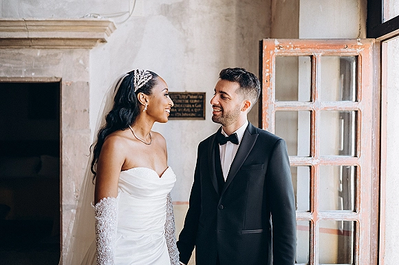 Couple portrait of bride and groom looking at each other, holding hands by a window, bride in strapless gown with veil and tiara