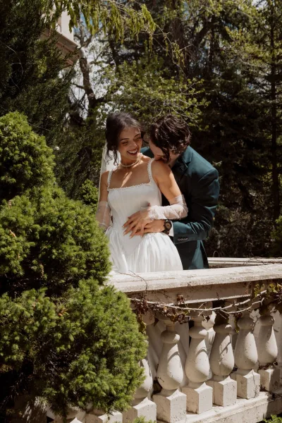Couple portrait of a bride and groom embrace on a stone balcony, her sheer gloves catching light amid vines and garden trees.