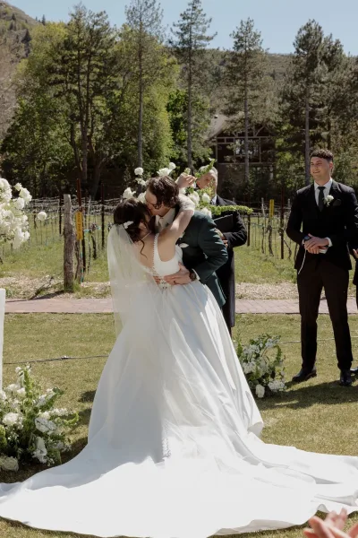 Wedding first kiss during an outdoor ceremony moment, the bride’s veil lifts as they embrace amid vineyard rows and a mountain hillside.