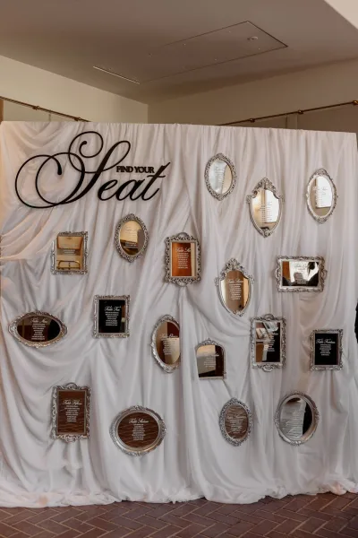 Wedding seating chart wall displayed on mirrors, in an indoor venue with a visible ceiling and brick floor