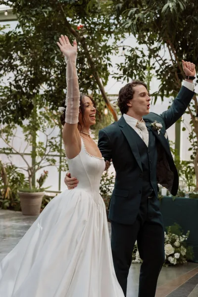 Wedding recessional captures a just married celebration as arm-in-arm newlyweds cheer in a greenhouse aisle, the bride in elegant bridal gloves.