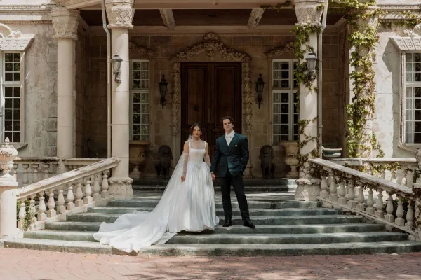 Couple portrait of the bride and groom holding hands on stone steps before an ivy-clad mansion, the bride’s long train cascading behind.