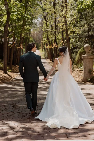 Couple portrait of a bride and groom walking hand in hand along a tree-lined brick path, her long veil trailing through the garden greenery.