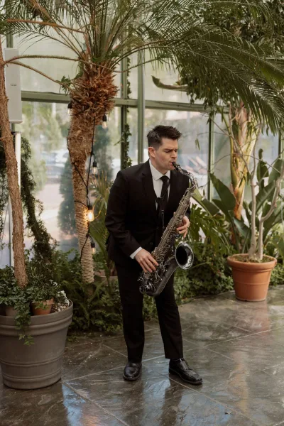 Wedding saxophonist delivering live wedding music in a glass-walled greenhouse, framed by tropical plants and soft string lights.